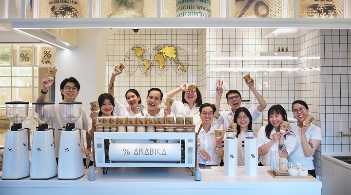 Group of % Arabica employees standing behind a coffee machine with their hands in the air