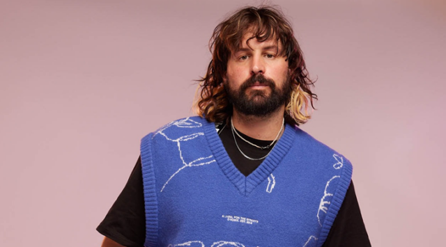 A portrait of Homie co-founder Marcus Crook wearing a blue knitted vest and black t-shirt against a mauve background