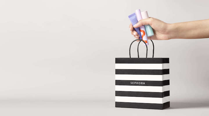 A hand clutching several beauty products above a Sephora shopping bag, set against a light gray background.