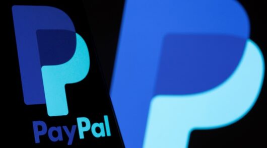 The PayPal logo is seen on a smartphone in front of the same logo displayed in this illustration